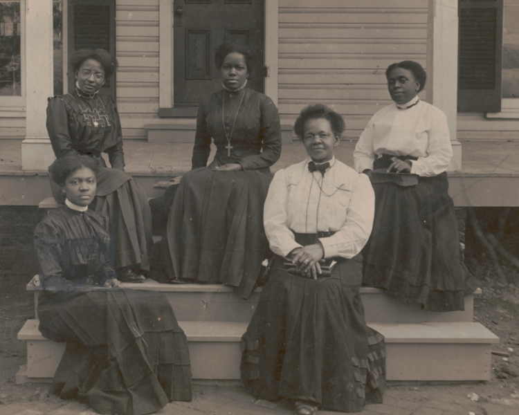Nannie Helen Burroughs sits with four other Black women in Victorian dress on the steps to the front porch of a building.