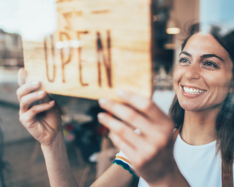 A woman is smiling as she hangs a wooden OPEN sign in a window