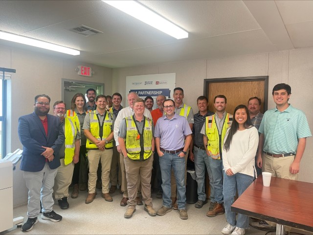 Joel Batiz, Area Director, Birmingham Area Office, Birmingham Region stands in a group inside the project worksite office, with 18 representatives from Brasfield & Gorrie after the signing ceremony.  