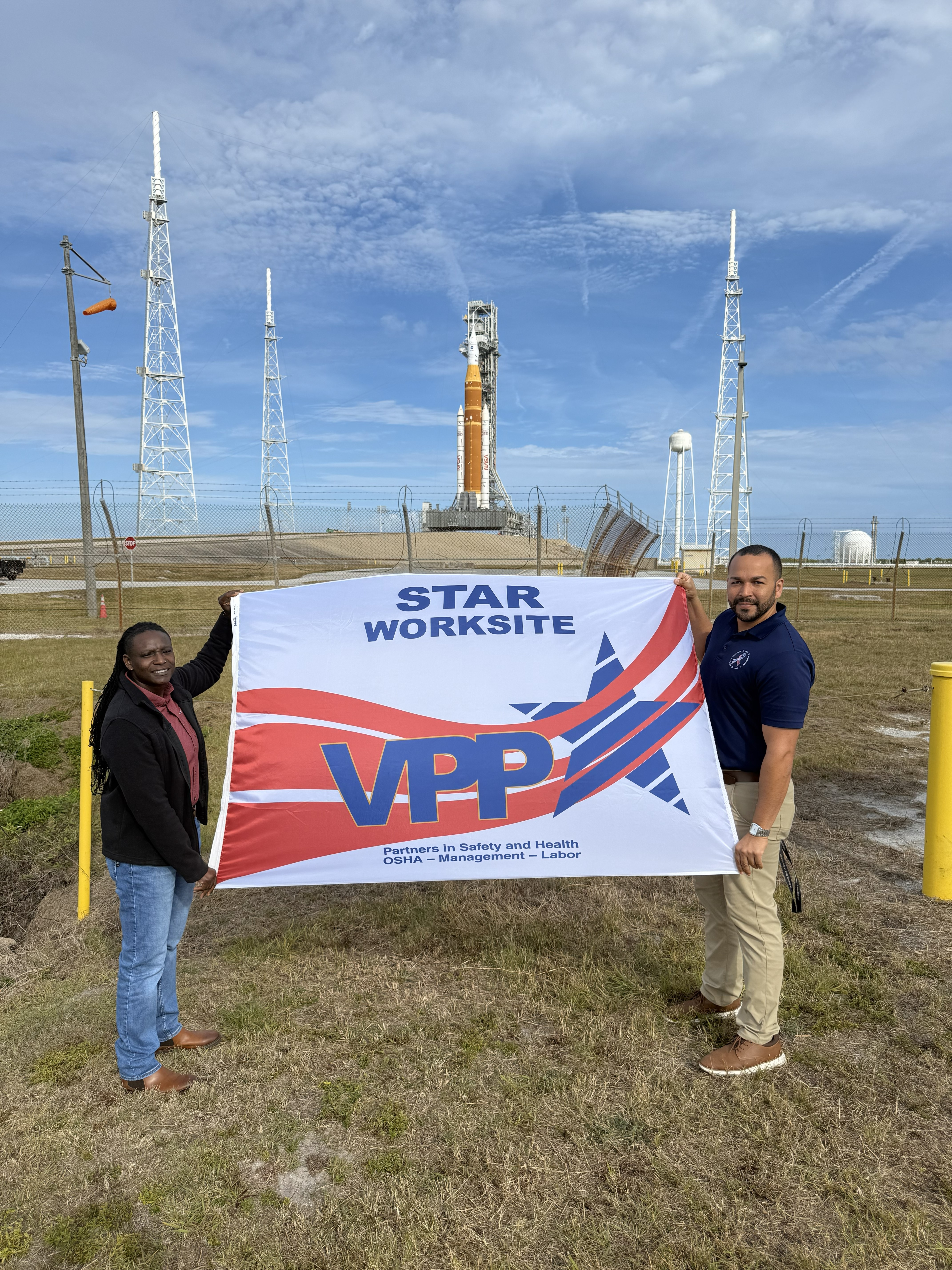 With the Artemis II mission rocket on its launchpad in the background, Tommiah Walker, Occupational Safety Specialist and Jeffrey Silva, VPP and Federal Safety Programs Manager, display the KSC VPP Star flag in the foreground.  (226 characters with spaces)
